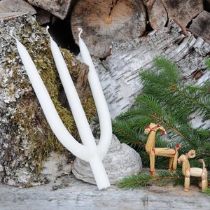 Puede incluir: Una vela blanca de tres mechas con un diseño único en forma de Y. La vela se coloca sobre una superficie de madera con corteza de abedul, musgo y ramas de hoja perenne. Dos decoraciones de renos de paja añaden un toque festivo.