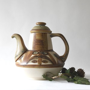 May include: A ceramic teapot with a lid, handle, and spout. The teapot features a cream-colored base, with brown and tan painted designs. The lid is olive green. The teapot is a decorative item.