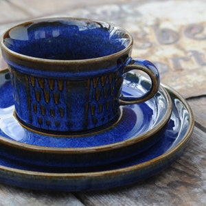 May include: A cobalt blue ceramic teacup and saucer set. The cup has a rounded shape with a handle and a textured pattern on the lower half. The set includes two additional plates, all with a dark brown trim.