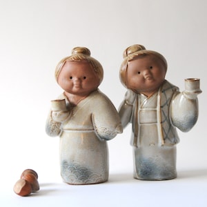 Może przedstawiać: Dwie ceramiczne figurki postaci w tradycyjnych strojach, każda trzymająca mały kubek. Figurki mają brązową skórę i są ubrane w kremowe szaty z niebieskimi wzorami fal na obszyciu. Jedna figurka ma złamaną głowę.