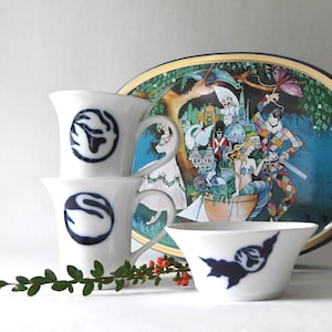 May include: Two white ceramic mugs and one white ceramic bowl, each featuring a dark blue swan logo. Behind them stands an oval tray decorated with a colorful illustration of a harlequin, a mermaid, a swan, and a soldier in a park setting.