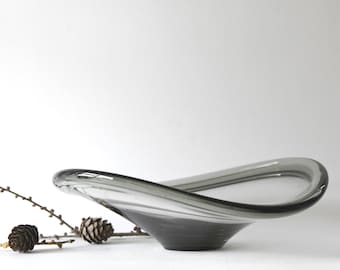 May include: A grey glass bowl with a wavy design. The bowl is shaped like a shallow dish with a wide, rounded rim and a slightly pointed base.