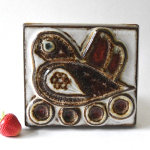 May include: Square ceramic tile featuring a bird design in brown, white, and red. The bird has a large eye and a stylized wing. The tile has a glossy finish and is likely a decorative piece.