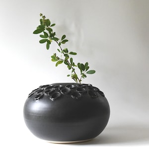May include: A matte black ceramic vase with a rounded, bulbous shape and a textured top surface featuring multiple small circular openings. A single green leafy branch with small buds is inserted into one of the openings of the dark vessel.