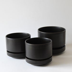May include: Three matte black cylindrical ceramic planters of varying sizes, each featuring a distinct horizontal groove near the base that separates the main body from the integrated saucer, creating a clean and uniform appearance.