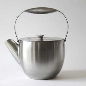 Op de afbeelding: Een zilveren theepot van roestvrij staal met een gebogen handvat en een deksel. De theepot heeft een gladde, glanzende afwerking.
