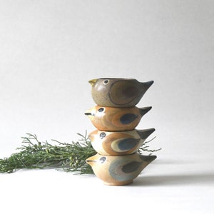 Puede incluir: Una pila de cuatro cuencos de cerámica con forma de pájaro. Los cuencos son de tonos marrón, beige y azul, con una textura moteada. Cada cuenco tiene un pico y una pequeña abertura en la parte superior. Los cuencos están apilados uno encima del otro.