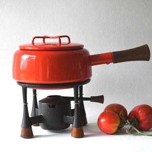 May include: A vintage fondue pot set with a bright red enamel pot and lid, a black metal stand, and a dark wooden handle. The set includes a black burner and is accompanied by three ripe tomatoes.