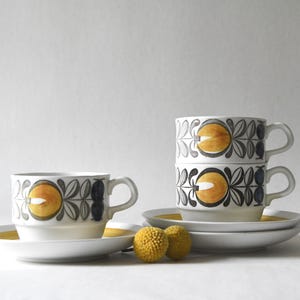 May include: Three white ceramic cups and saucers featuring a repeating pattern of yellow circular fruit and dark grey leaves. One cup stands alone on a saucer, while two cups are stacked on a second saucer. Two small yellow spherical flowers sit in front.
