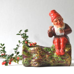 Può includere: Una fioriera a forma di gnomo in ceramica, con cappello e pantaloni rossi, giacca marrone e barba bianca. Lo gnomo è seduto su una fioriera a forma di tronco con dettagli strutturati verdi e marroni. Un rametto di verde con bacche rosse è a sinistra.