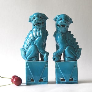 May include: Two turquoise ceramic Foo Dog statues, each perched on a square base. The dogs have detailed features and are positioned side-by-side. Two red cherries are in the foreground.