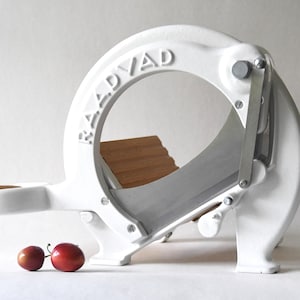 Puede incluir: Cortadora de alimentos de hierro fundido blanco con base de madera y cuchilla plateada. La cortadora está etiquetada como "RADVAD". Dos ciruelas rojas están en primer plano.