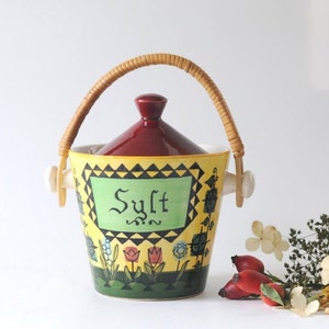 Puede incluir: Un recipiente de sal de cerámica amarillo y rojo con un asa de mimbre. El recipiente tiene una etiqueta verde que dice "Sal" y está decorado con flores y un patrón geométrico en blanco y negro.