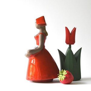 Puede incluir: Figura de cerámica de una mujer con vestido y sombrero rojos, sosteniendo una cesta. Un tulipán rojo y una fresa están junto a la figura. El tulipán tiene hojas verdes. El fondo es blanco.