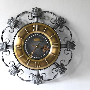 Op de afbeelding: Vintage wandklok met een goudkleurige wijzerplaat en Romeinse cijfers. De klok heeft een donkergrijze kern en een sierlijk frame in smeedijzeren stijl. De wijzers zijn koperkleurig. Op de wijzerplaat staat de merknaam "Bisch".
