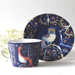 Könnte beinhalten: Ein marineblaues Keramik-Tassen- und Untertassen-Set. Die Tasse und die Untertasse zeigen ein skurriles Walddesign mit einem roten Vogel und einer Eule. Das Set hat eine weiße Innenseite und goldene Akzente.
