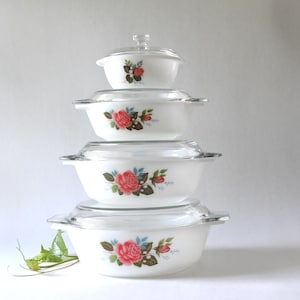 Peut inclure: Une pile de quatre plats à four en verre opalin blanc avec des couvercles en verre transparent. Chaque plat présente un motif floral de roses roses avec des feuilles vertes et des accents bleus. Les plats sont empilés par taille décroissante.