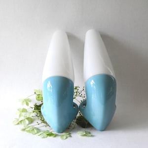 May include: Two vintage wall sconces. Each sconce has a white, elongated, teardrop-shaped glass shade above a light blue, curved ceramic base. The sconces are set against a white background with green foliage.