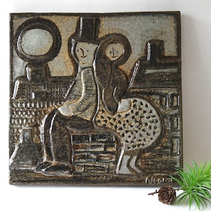 Op de afbeelding: Vierkante keramische wandkunst met een stel in een stedelijke omgeving. Het kunstwerk is in bruin-, grijs- en wittinten, met een getextureerd oppervlak. De man draagt een hoge hoed en de vrouw heeft een polkadotjurk. Het stuk is gesigneerd "Noora".