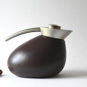 May include: A modern, brown ceramic coffee pot with a stainless steel handle and lid. The pot has a rounded, egg-like shape. Two hazelnuts are in the foreground.