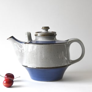 Peut inclure: Théière en céramique avec une base bleue et un corps gris moucheté. La théière a un couvercle assorti et une anse incurvée. Deux cerises sont au premier plan. La théière mesure environ 20 cm de haut.