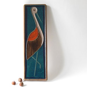 May include: Rectangular ceramic wall art with a stylized bird design. The bird has a white head and neck, an orange chest, and brown striped wings. The background is teal, framed by a brown border. A decorative piece for any home.