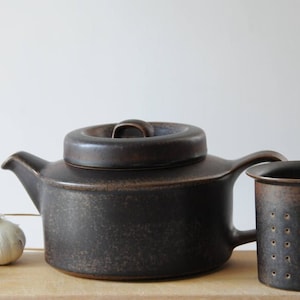 Può includere: Una teiera in ceramica marrone scuro con un coperchio e un filtro per il tè in ceramica marrone abbinato. La teiera ha una finitura macchiata.
