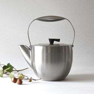 以下が含まれることがあります： 黒い取っ手と蓋が付いた銀色のステンレス製のティーポット。ティーポットは丸みを帯びたボディと側面に注ぎ口があります。ティーポットは白い表面の上に置かれています。