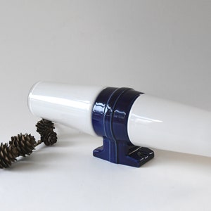 May include: White ceramic vase with a blue band around the middle. The vase is lying on its side on a white surface.