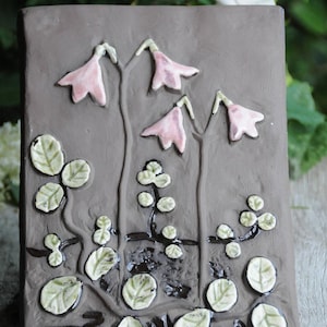 May include: A brown ceramic tile featuring a raised design of pink flowers and green leaves. The tile has a rustic, handmade appearance.