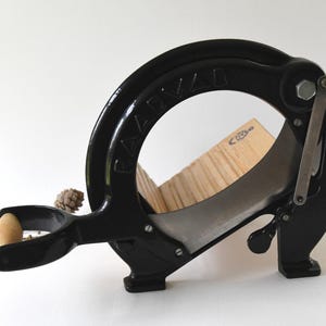 Puede incluir: Cortador de pan de hierro fundido negro con una tabla de cortar de madera y un mango de madera. El cortador está etiquetado como "RAZIVAD".