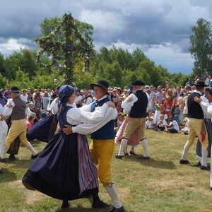 Puede incluir: Un grupo de personas con trajes folcl&oacute;ricos tradicionales bailan al aire libre. Los hombres visten chalecos, sombreros y pantalones amarillos o beiges. Las mujeres llevan faldas largas y pa&ntilde;uelos en la cabeza. Un poste decorado se alza al fondo, y una multitud observa.