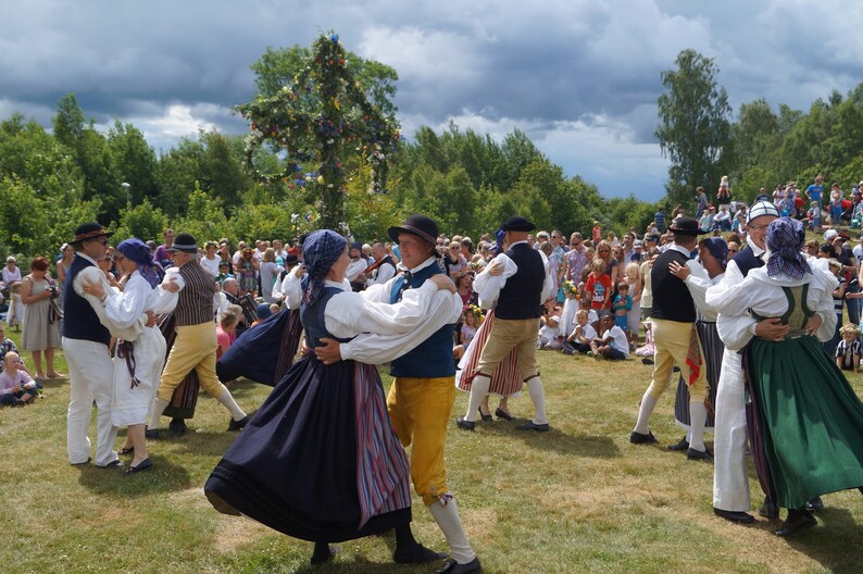 Puede incluir: Un grupo de personas con trajes folcl&oacute;ricos tradicionales bailan al aire libre. Los hombres visten chalecos, sombreros y pantalones amarillos o beiges. Las mujeres llevan faldas largas y pa&ntilde;uelos en la cabeza. Un poste decorado se alza al fondo, y una multitud observa.