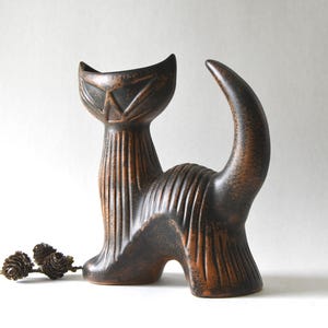 Puede incluir: Una figura de gato de cerámica de color marrón oscuro con un cuerpo texturizado y acanalado y una cara angular estilizada. El gato está representado en posición de pie con la cola curvada hacia arriba. Dos pequeñas piñas marrones están cerca.