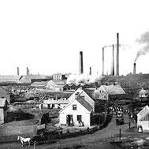 May include: Black and white photograph of a factory with tall smokestacks and several buildings. The factory is surrounded by smaller houses and a dirt road.