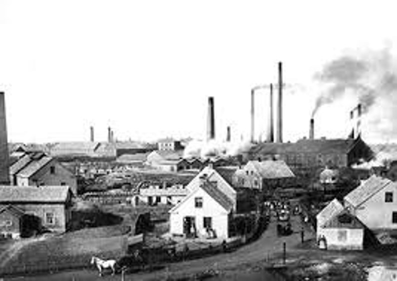 May include: Black and white photograph of a factory with tall smokestacks and several buildings. The factory is surrounded by smaller houses and a dirt road.