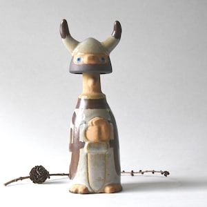 May include: Ceramic figurine of a Viking with a horned helmet, blue eyes, and a brown and white tunic. The figurine has a stylized design and is standing upright. A small pine cone and twig are in the foreground.