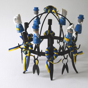 Puede incluir: Un candelabro de metal negro con m&uacute;ltiples portavelas, adornado con cintas azules y amarillas y peque&ntilde;as figuras de madera vestidas con trajes tradicionales suecos. Las figuras est&aacute;n pintadas con detalles intrincados.