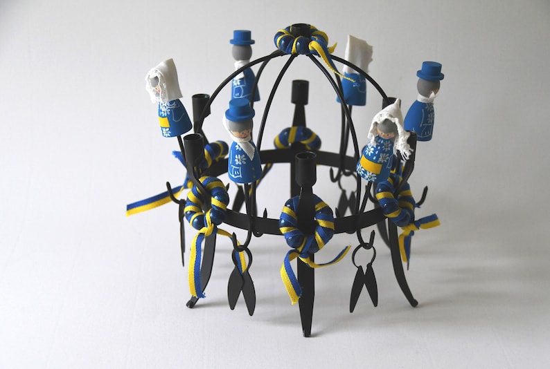Puede incluir: Un candelabro de metal negro con m&uacute;ltiples portavelas, adornado con cintas azules y amarillas y peque&ntilde;as figuras de madera vestidas con trajes tradicionales suecos. Las figuras est&aacute;n pintadas con detalles intrincados.