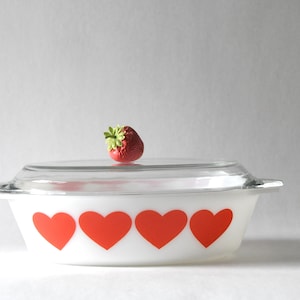 May include: White casserole dish with a clear glass lid and red heart designs. A red strawberry with green leaves sits atop the lid. The dish has two handles and is likely used for baking or serving.