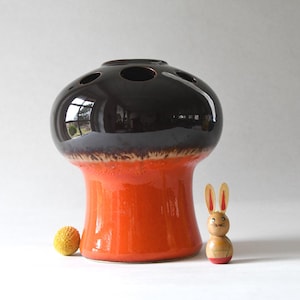 May include: A ceramic vase with a glossy black top, gray midsection, and orange base. The vase has several holes on top. A small wooden bunny and a yellow ball are next to the vase.