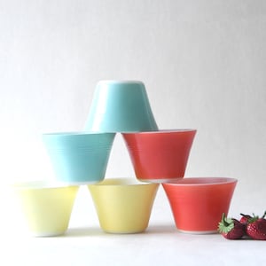 Könnte beinhalten: Sechs bunte Vintage-Glasschalen in Pyramidenform gestapelt. Die Farben sind Aqua, Rot und Gelb. Frische Erdbeeren liegen rechts. Auf weißem Hintergrund.