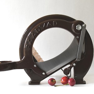 Puede incluir: Un deshuesador de cerezas de hierro fundido marrón con un mango de madera y una cuchilla plateada. El deshuesador está sobre una superficie blanca con tres cerezas rojas delante.