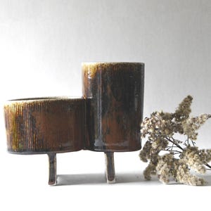 May include: A brown ceramic planter with a textured surface and three legs. The planter has two connected sections, one round and one cylindrical. A dried floral arrangement is placed to the right of the planter.