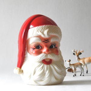 May include: A molded plastic Santa Claus head figurine featuring a bright red hat with a white pom-pom, a white beard, and painted facial features including blue eyes with long black eyelashes, rosy cheeks, and a red mouth, accompanied by two deer.