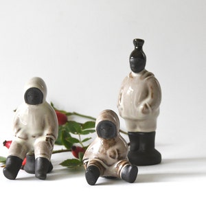May include: Three ceramic figurines of people wearing hooded coats. The figurines are in a seated position and have black faces. The figurines are on a white background with a sprig of red berries.
