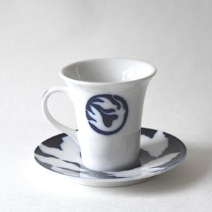 May include: A white ceramic cup and saucer set. The cup features a dark blue circular logo with a stylized flame or leaf design on the front. The matching saucer is decorated with a dark blue pattern of large, overlapping leaf shapes.