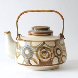 Op de afbeelding: Een keramische theepot met een crèmekleurige basis, versierd met bruine, blauwe en bruine cirkelvormige patronen. Het heeft een tuit, een deksel en een geweven handvat. De theepot is ontworpen voor het zetten van thee.