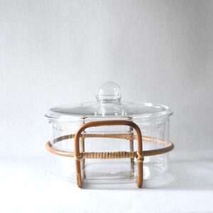 May include: A clear glass casserole dish with a matching glass lid featuring a spherical knob handle. The dish is encased in a light brown, curved rattan frame that wraps around the sides and forms two integrated handles on opposite sides.