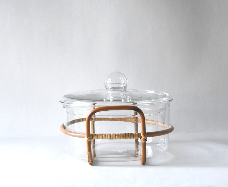 May include: A clear glass casserole dish with a matching glass lid featuring a spherical knob handle. The dish is encased in a light brown, curved rattan frame that wraps around the sides and forms two integrated handles on opposite sides.